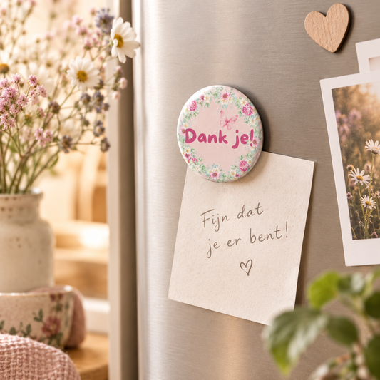 Een ronde fotomagneet van 38mm met de tekst "Dank je!" op een koelkast, die een kaartje met "Fijn dat je er bent!" vasthoudt, omringd door een sfeervol interieur met een bos bloemen, een kleedje en een plant.