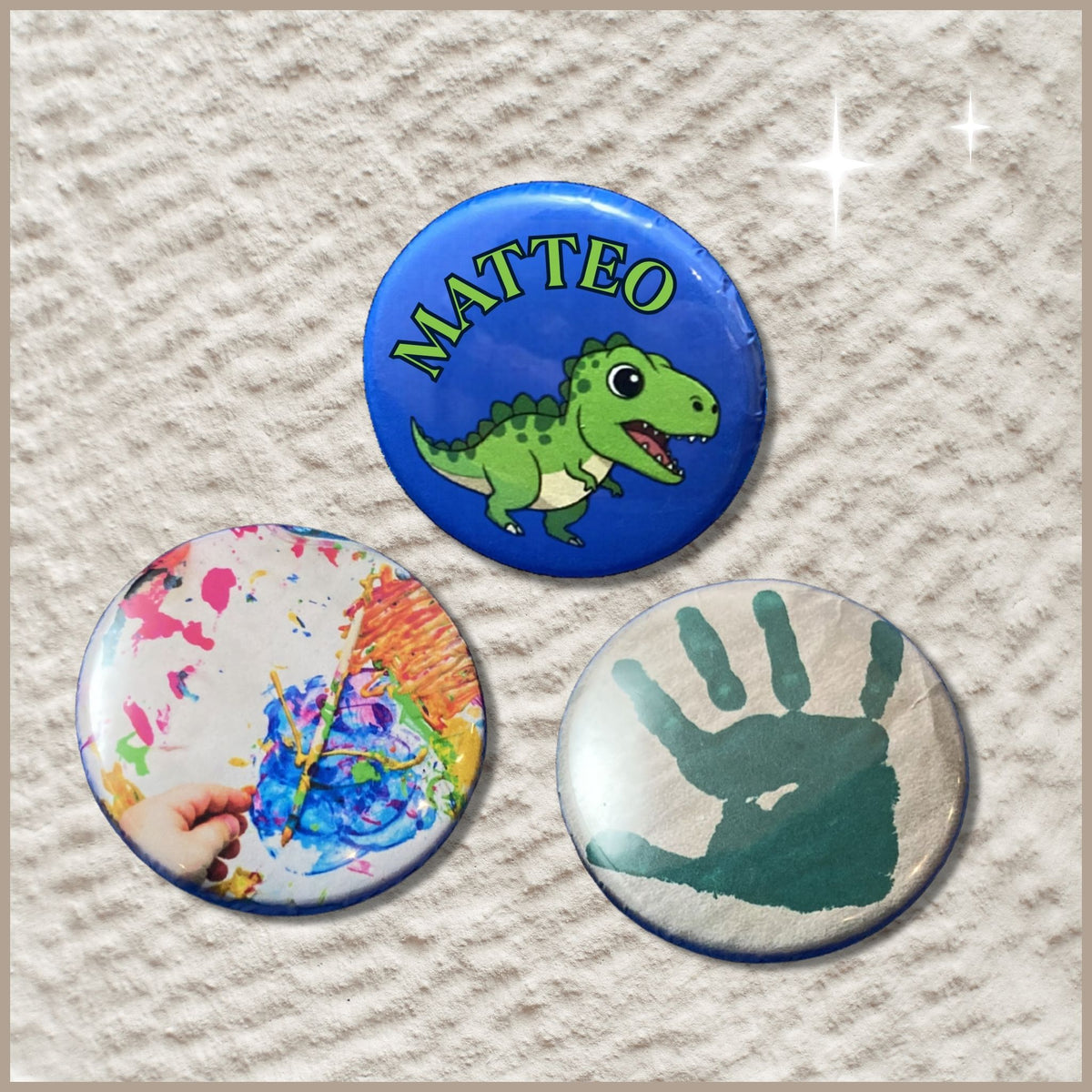 Een set van drie ronde buttons van 38 mm, bestaande uit een button met een verfafdruk van een kinderhandje, een button met een kleurrijke kindertekening en een naambutton met een dinosaurus-illustratie.
