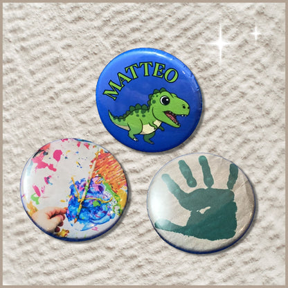 Een set van drie ronde buttons van 38 mm, bestaande uit een button met een verfafdruk van een kinderhandje, een button met een kleurrijke kindertekening en een naambutton met een dinosaurus-illustratie.
